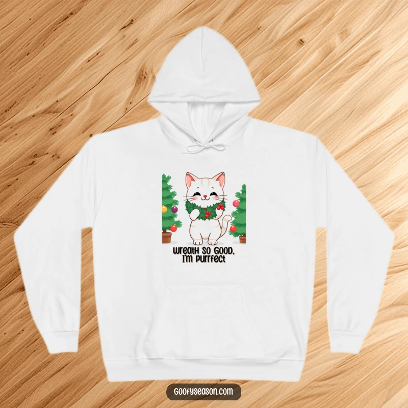 Funny hoodie depicting a cheerful cat in a wreath, placing the last ornament on a beautifully decorated Christmas tree.