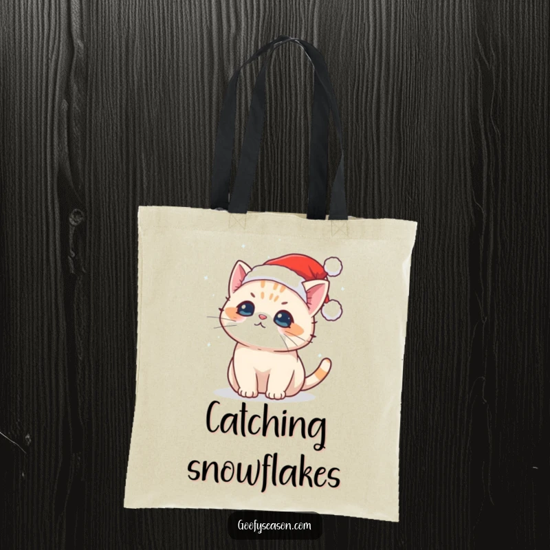 Funny tote bag featuring a cute Kawaii cat in a Santa hat catching falling snowflakes, a charming holiday accessory.